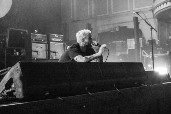 Black and white landscape photo of a bearded musician sat on the stage and leaning over the riser speakers.