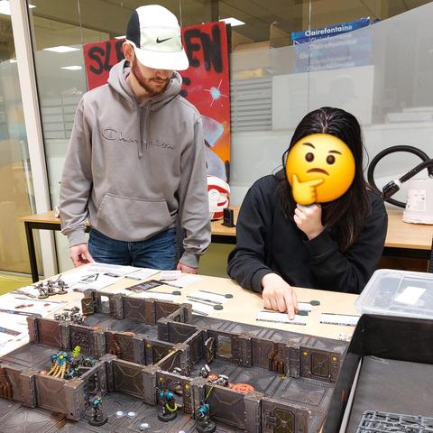 One student explaining kill team to another one. The latter is scratching her chin. There appears to be epidemic of Chin Itch going on.