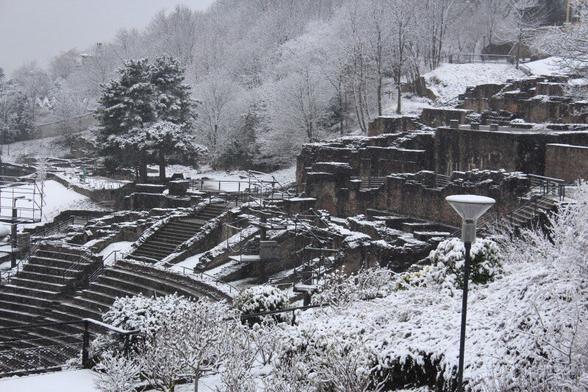 A serene winter scene featuring the ancient Roman theater of Fourvière in Lyon, France, blanketed in a layer of pristine snow. The historic stone structure, with its semi-circular seating and stage area, stands out against the white landscape, offering a unique blend of ancient architecture and seasonal beauty.