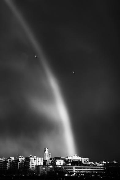 black and white. portrait format. glowing rainbow in the dark sky above the frankfurt university hospital. two seagulls cross the scene.