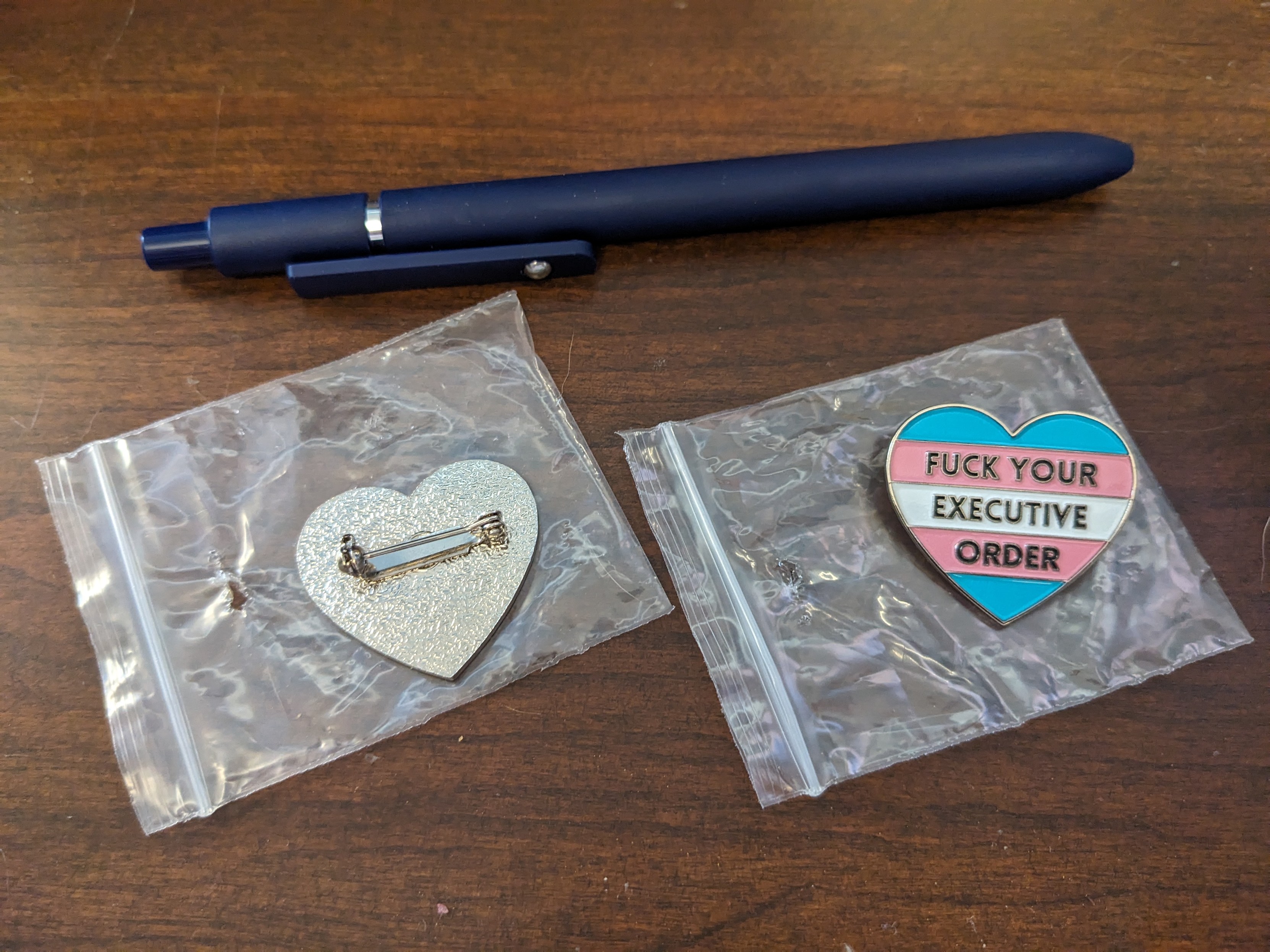 A small heart-shaped pin stylized with the Trans Flag background, that says "FUCK YOUR EXECUTIVE ORDER" in black block-letters. The pin is "soft enamel" printed on metal, with a safety-pin backing. There is a pen for scale, these are about 1.25 inches in size.