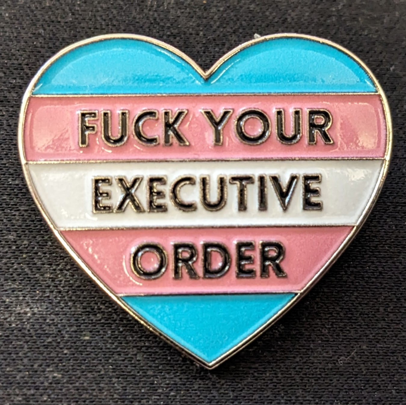 A close-up of the front of the pin. It is a metal pin in a heart shape, stylized with the trans flag as a background, and says "FUCK YOUR EXECUTIVE ORDER" in large black block-letters along the three center stripes.