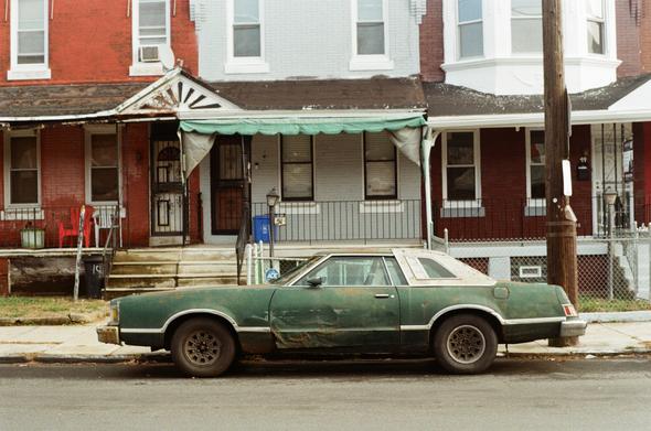 A very worn out late 1970’s green Mercury Cougar Coupe with a white half landau vinyl top sits in front of a brick red, grey washed and burgundy row home in the Cobbs Creek Neighborhood of Philadelphia. The car has various damage and rust spots, especially on the drivers door.