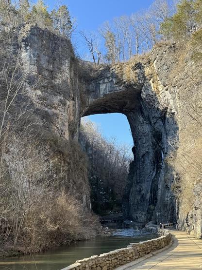 This photo was taken at about 2pm on Monday, and the sun is only slanting in on about half of the gully formed by Cedar Creek.  It is amazing what water can do to limestone... given several hundred thousand years.

The photo shows both sides of a steep gorge, with a small creek (about 30' across?) in the center, and a pathway edged in a low rock wall on the right, curving down to pass under the bridge itself.

The structure of the bridge looks to be also about 30' thick, so from where this was taken you can see blue sky both above and below it.  The length is probably a similar dimension.  I don't think people are allowed to walk on it, which is a very good thing.  There are a few small trees growing on the bridge itself, as there are on both top sides of the cliffs.

It looks a little like a very large dumbbell.