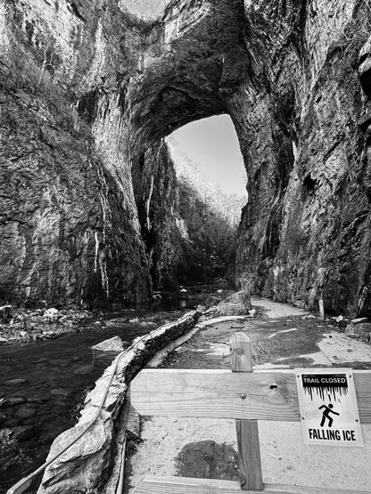 Closer to the bridge and in black and white, this photo captures some of the icicles and sheets of ice coating the walls of the cliff under the bridge itself.  

The photo is taken directly in front of a sawhorse blocking the trail forward that would go under the structure.  On the blockade is a sign which says "Trail Closed, Falling Ice" and shows a stick figure person struggling to walk, and about to be impaled from above by a row of huge ice daggers.

Good idea, we stopped right here.  Many pieces of ice fell and shattered on the trail and rocks during the 20 minutes we were enjoying the area.