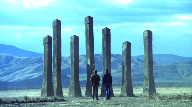 A still from Phase IV, two men in the desert look at strange rectangular ventilation towers the only above ground evidence of the massive nest built by the ants who have started to cooperate across species. The towers are three times the height of a man and like brutalist architecture.