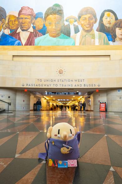 Photo of the Creature Mastodon stuffed toy sitting on the floor facing of Patsaouras Transit Plaza at Union Station Los Angeles. The Creature is in its purple tote in the middle of the Union Station Walkway beneath the “City of Dreams/River of History” mural, facing the camera with its back turned toward the train platforms. At the base of its bag are three TAP cards: the Black History Month 2025 Commemorative TAP card from LA Metro, the default TAP card, and the Valentines Day 2025 Commemorative TAP card from LA Metro.