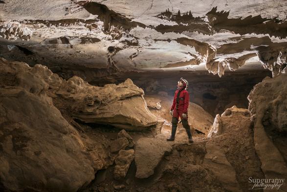 A person in a red oversuit looks up towards ceiling formations. The rock is yellow-brown, covered in dust and mud, while the ceiling looks cleaner in color. A warm back lit scene.