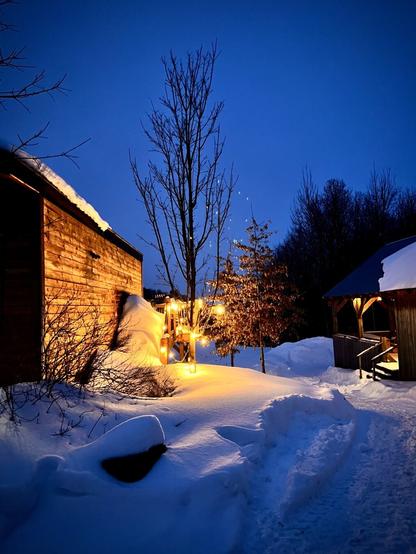 A winter evening scene with a snow-covered landscape. A wooden cabin on the left emits a warm glow from its exterior lighting. A snow-covered pathway leads toward a wooden gazebo on the right. A leafless tree stands in the center, with a smaller tree nearby decorated with glowing string lights. The deep blue twilight sky contrasts with the warm yellow-orange tones of the artificial lights.