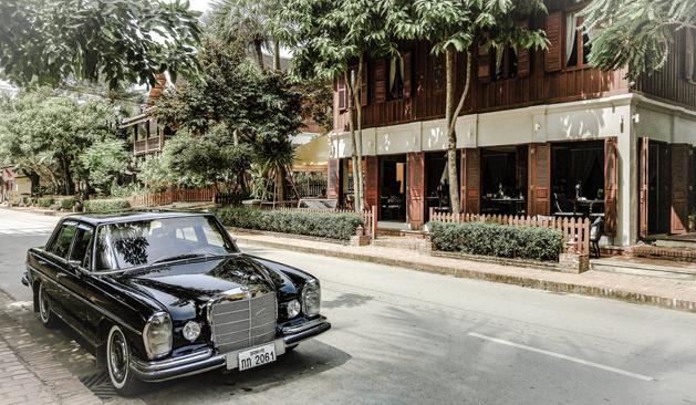 Photo shows an old, classic black Mercedes Benz car from the 1950s parked at the street in front of a building of typical colonial architecture. The building is in red bricks with white and brown details. It is framed by green trees and hedgerows.