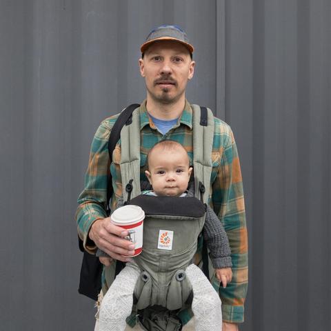 Man with a baby in a carrier on his chest. He is wearing a hat, has a bag on his shoulder, and is holding a coffe cup.