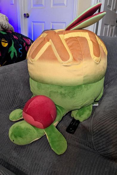 A lifesize Appletun plush is sitting on an armchair with an Amazon Fire Cube remote under it's front paw. The creature is a dragon with apple pie motifs.