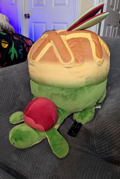 A lifesize Appletun plush is sitting on an armchair with an Amazon Fire Cube remote under it's front paw. The creature is a dragon with apple pie motifs.