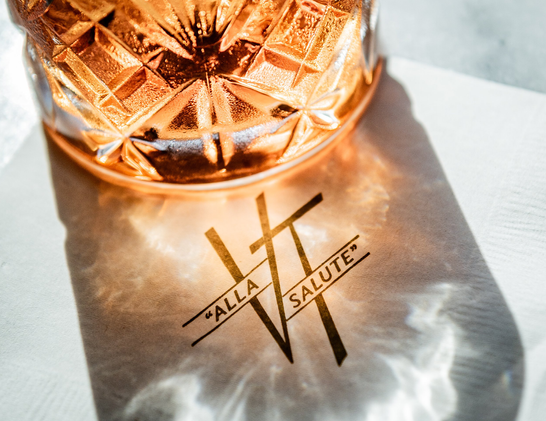 Photo of a cocktail glass with shadow and reflection on a printed surface reading VT: “Alla Salute”.