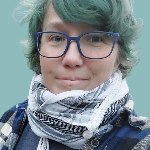 Foto af Ivy-Oak, en hvid nonbinær person, med grønt hår og blå briller. De har et hvidt og sort kuffiyeh om halsen, og en grå, blå og hvid flannel skjorte på. Baggrunden er lyseblå