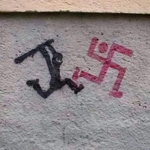Stencil street art on a concrete wall depicting a black figure, possibly a representation of resistance or justice, wielding a club and chasing a red swastika-like figure that appears to be running away. The artwork conveys a strong anti-fascist message, symbolizing the fight against Nazism and hate ideologies.