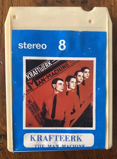 A photograph of a vintage Italian white plastic 8-track tape entitled "The Man Machine" by Kraftwerk. Inset cover photo shows four impeccably-groomed band members wearing matching black ties and red shirts looking off to the right. The pose is reminiscent of the social realist art encouraged in post-WWII Soviet Bloc nations. Text at the top reads "stereo 8". Text at bottom reads "KRAFTEERK / THE MAN MACHINE".