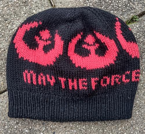 Schwarze Mütze mit rotem Einstrickmuster, das Rebellen Logo aus Star Wars und dem Schriftzug "May the Force"