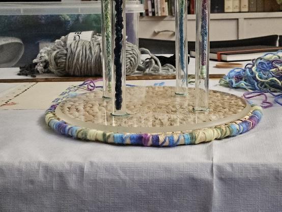 In the foreground, there is a round placemat made of straw and cane; the edges of it have been wrapped with colourful yarn. Sitting on top of the placemat is a THING with four transparent plastic legs attached to a round transparent plastic base. In the background you can see a large needle with the colourful yarn attached to it; behind that is a ruler, behind that are balls of yarn.
