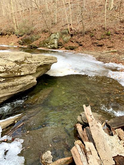 The water flowing from the right side moves swiftly along the big boulder, remaining unfrozen. But where the current slows, a layer of ice forms on the surface. The water has a green hue.