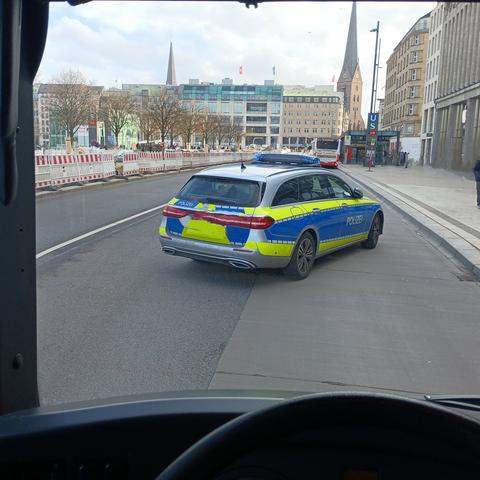 Ein Foto, aufgenommen aus dem Inneren eines Fahrzeugs, zeigt einen blauen und gelben deutschen Polizeiwagen, der auf einer Straße in einer Stadt steht. Im Hintergrund sind Gebäude, darunter Kirchtürme, und ein Bus zu sehen. Der Himmel ist bewölkt. Im Vordergrund ist ein Teil des Lenkrads des aufnehmenden Fahrzeugs sichtbar.