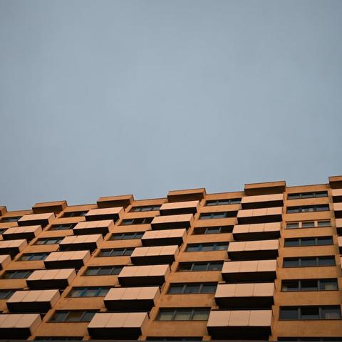 This is an image of a modern apartment building captured from a low-angle perspective against a clear sky. The building has a repetitive pattern of rectangular balconies or sunshades, creating a geometric aesthetic. The facade appears to be a mix of warm tones, possibly orange or beige, with dark windows. The image composition emphasizes the height and structure of the building.