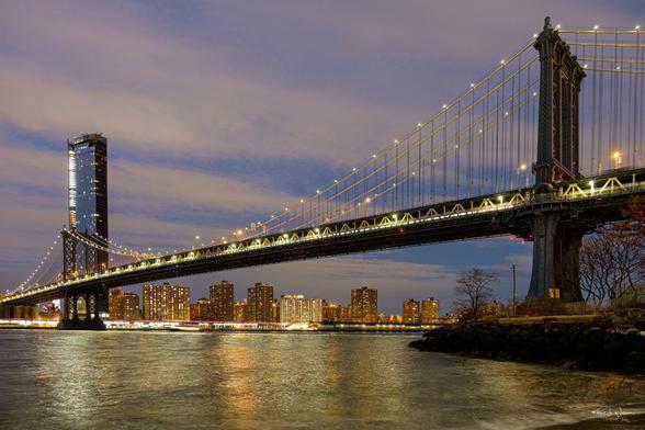 Night view of the Manhattan Bridge in New York City, with glowing lights and reflections on the East River.