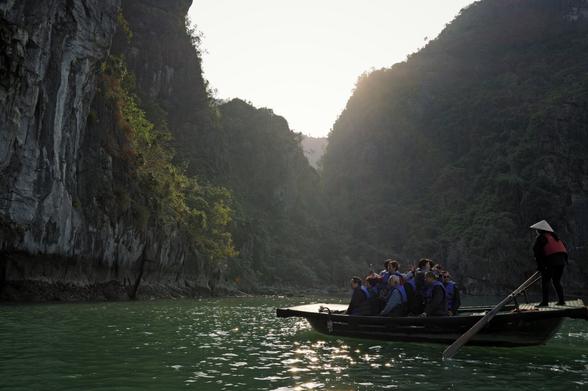 Ein traditionelles Ruderboot mit Touristen, die mit Handys in alle Richtungen fotografieren, fährt durch die Halong-Bucht in Vietnam. Die Passagiere tragen Rettungswesten, während eine Bootsführerin mit einem Non-La-Hut am Heck steht und das Boot mit einem Ruder steuert. Steile Kalksteinfelsen, bewachsen mit dichter Vegetation, ragen links und rechts aus dem smaragdgrünen Wasser. Die tief stehende Sonne wirft ein warmes Licht auf die Szenerie.