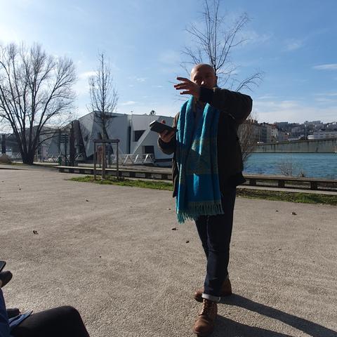 Photo d'Olivier debout, à contre-jour, il porte une large écharpe bleue clair autour du cou. En arrière plan, on voit le Rhône et un grand bâtiment blanc (le théâtre)