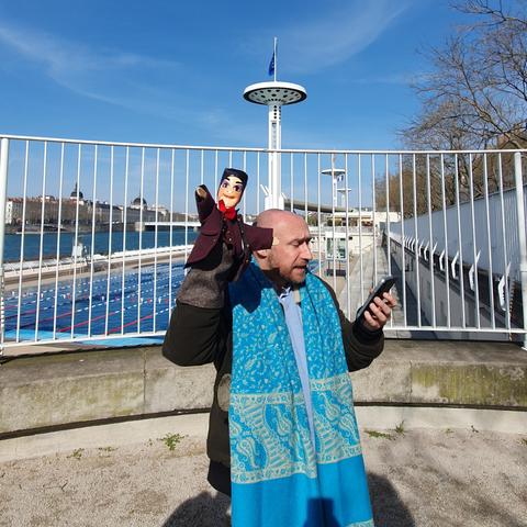 Photo d'Olivier debout, il porte une large écharpe bleue clair autour du cou et dans sa main droite il tient Guignol. Dans sa main gauche il tient son portable et il lit. En arrière plan, on voit la piscine du Rhône avec ses lignes d'eau et l'un de ses 4 grands pylones.