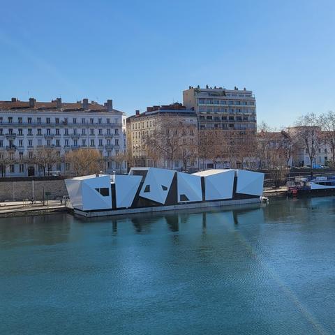 Photo du théâtre de l'île Ô. C'est un bâtiment flottant blanc composé de sortes de gros cubes blancs. La photo est prise depuis l'autre côté du Rhône