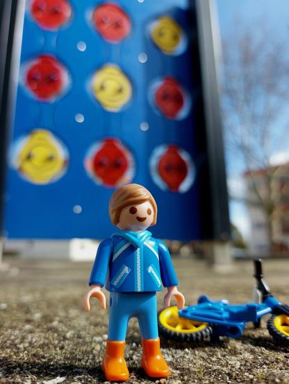 Criança playmobil vestida de azul, cabelo castanho claro, com sorriso e bicicleta azul bmx deitada atrás. Em background o jogo do galo / jogo da velha / tic tac toe