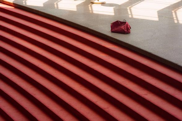 The image shows a set of red carpeted stairs leading up to a platform. On the platform, there is a red bag placed. The lighting creates shadows on the floor, adding pattern and depth to the scene.