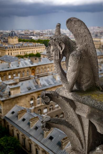 Color photo of one of the “grotesques” sculptures perched on the balustrade of the cathedral's facade, hand on chin, gazing out over the buildings of Paris below.