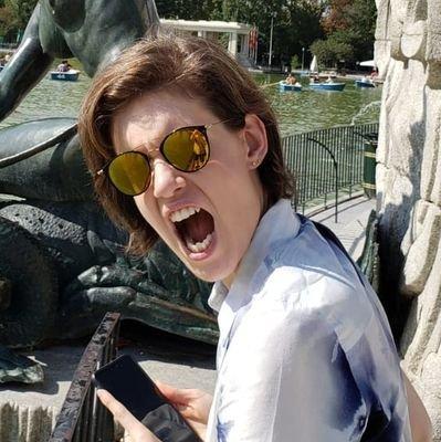 Head-and-shoulders color photo of author Shelley Lavigne, a fair-skinned person with red-brown hair worn in a one-length bob at chin length. They are wearing a white shirt and large sunglasses with gold lenses. They appear to be sitting by a pool and a bronze statue. They look up from a smartphone, over their shoulder, into the camera, screaming ferociously.