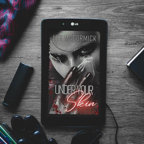 under your skin by lee mccormick ebook cover, showing a face taking off a transparent plastic face shield, dark red staining their fingers, and blood spatter on the edges of the cover. black framed lg tablet is placed on a dark grey wooden table, with various dark items strewn around, such as a lighter, leather gloves, round black shades, face mask.