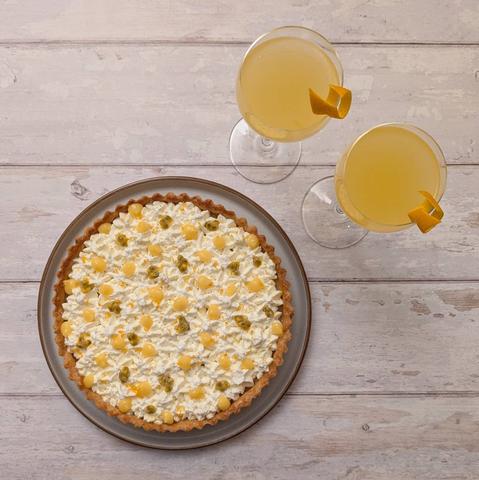 A square photo from above of a tart sitting on a plate next to two cocktails. The tart has a golden shortcrust case and a whipped-cream topping made up of lots of stars piped closely together. This is decorated with some passion-fruit seeds and wee blobs of yellow passion-fruit curd, then sprinkled all over with some orange zest. The cocktails are two light-orange translucent drinks in Nick and Nora glasses with orange-peel garnish on the glass. They all sit on a white wooden background.