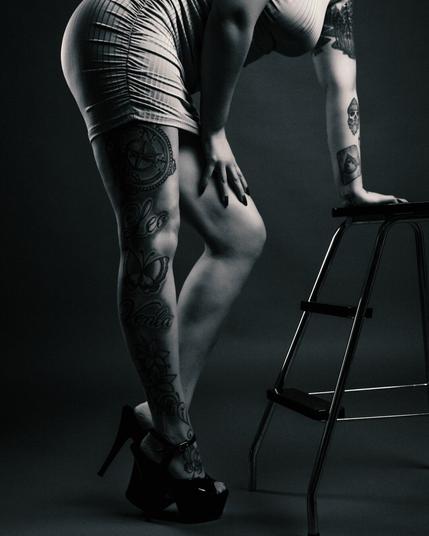 A black and white studio portrait of a tattooed woman wearing a tight dress and high heels, leaning against a metal stool. The composition highlights her leg tattoos, including a compass, butterflies, and script, with a dramatic lighting setup emphasizing the contrast.