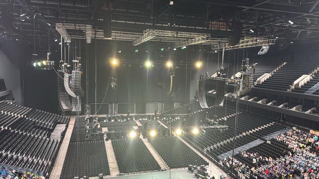 The image shows the interior of a concert venue with empty seating arranged in rows. A stage setup is visible, including suspended lighting and sound equipment overhead. The area appears ready for an upcoming event, with a few people seated in the foreground.