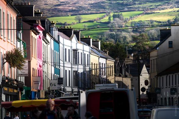 A street scene in Ireland featuring colourful buildings. The image shows a street with buildings painted in various colours such as pink, blue, and white. There are signs for 'tonic,' 'massage,' 'yoga,' 'bantry yarns,' and 'Fish Kitchen.' Hills are visible in the background.