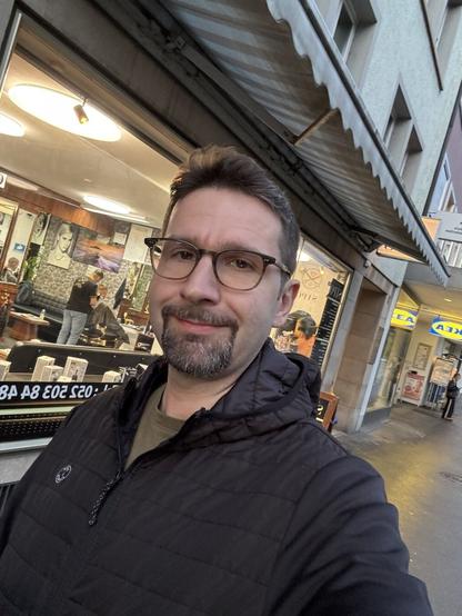 A person wearing glasses and a black jacket stands outside a shop with a barber pole and bright window display, smiling at the camera.