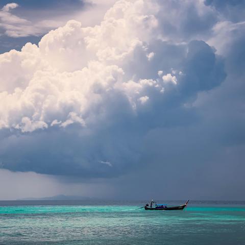 The image shows a scenic view of a large cloud formation over an ocean or sea. There's a small boat on the water, with a person standing in it. The water is a clear turquoise color, creating a contrast with the darker sky and clouds above.