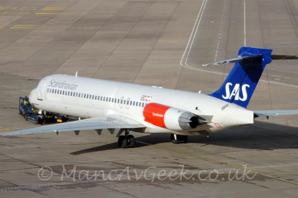 Rear side view of a white, twin engined jet airliner with the engines mounted on the rear fuselage, facing to the left but being pushed back to the right by a small blue tug attached to the nose-wheel.
The plane has silver "Scandinavian" titles on the upper forward fuselage, with grey "Airlines" titles on the lower fuselage.
The tail is a deep blue, with large white "SAS" titles at the base.
The engine pods are a bright red, with white "Scandinavian" titles.
Grey concrete apron fills the frame, with white road markings on the right, and yellow markings on the left.