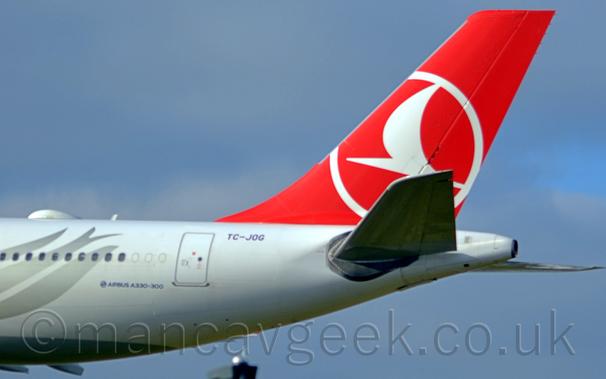 Closeup of the white rear fuselage and red tail of a jet airliner flying from right to left at a very low altitude.
The rear fuselage has a silver flower head on the rear fuselage, while the red tail has a stylised white flying bird inside the outline of a white circle.
Blue sky with hazy grey cloud fills the rest of the frame.