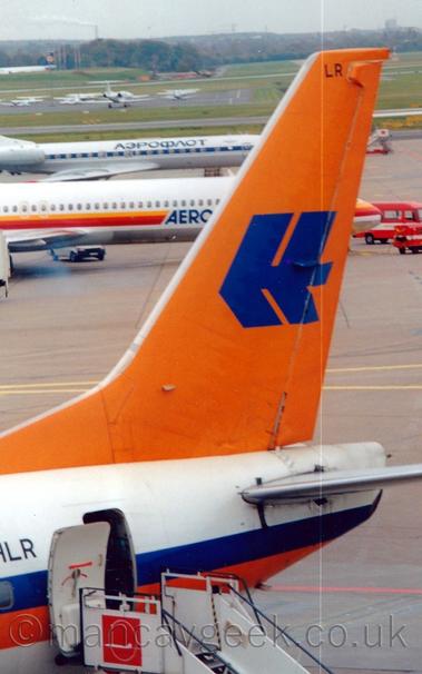 Closeup of the white rear fuselage and bright orange tail of a jet airliner parked facing to the left.
There is an orange and blue stripe running along the body, and  a stylised upper-case letter "H" in blue on the orange tail.
A set of white mobile airstairs is connected to the open rear door.
A pair of white jet airliners can be seen in the background on the left of the frame, with light aircraft and bizjets parked on an apron surrounded by grass further back, under cloudy grey sky.