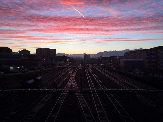 Une dizaine de voies de chemin de fer en parallèle partant de sous la position du photographe et se dirigeant vers le centre de l’image. On peut distinguer les quais de la gare de Renens au fond. Le soleil n’est pas encore visible, mais les couleurs du ciel indiquent qu’il va bientôt se lever. Les stratus dans le ciel reçoivent eux déjà la lumière du soleil par dessous, qui leur donne une couleur rose. Les voies de chemin de fer, la gare et la ville autour sont encore toutes dans la pénombre. Mais les rails sont mis en évidence car ils reflètent la lumière du ciel.