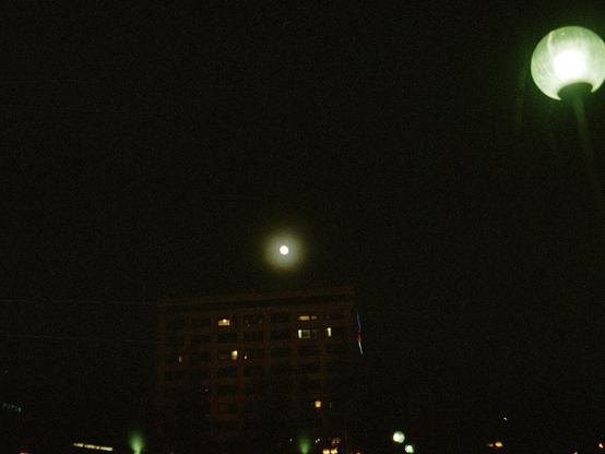 Full moon looming over a large apartment building in central Oulu. Street lights can be seen below, with one random park light post on the right side of the image.