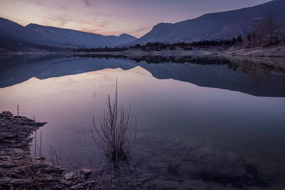 In the foreground you can see the calm waters of a reservoir. In the background, some mountains. Everything is bathed in a purple evening light.