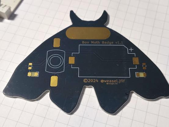 A close-up, slightly angled shot of a moth-shaped circuit board on a white grid background. The board is dark gray with gold-colored conductive traces. The top of the moth's head has two small, horn-like protrusions. The body of the moth has a rectangular gold area near the top, followed by a series of white lines forming a simple circuit diagram. Below the diagram, there is a copyright symbol, the year '2024', and the text '@weasel.jfif' and '@rough.rat'. The wings of the moth have small gold squares and circles at various points, indicating connection points. A small white object is visible in the upper left corner, slightly out of focus.