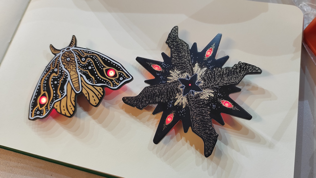 The photo shows two assembled PCB (Printed Circuit Board) art pieces lying on an open book with cream-colored pages. On the left is a moth-shaped PCB with visible electronic traces and components. The board is dark with yellow and white patterns resembling abstract blobs and lines. The moth's eyes are illuminated with red LEDs. On the right is an irregular star-shaped PCB with sharp edges, also showing visible electronic traces and components. It's dark with gold patterns and has red LEDs at several points, resembling eyes. Both PCB art pieces lie on the cream-colored background of the book's pages, and a fragment of an orange object is visible in the upper right corner.